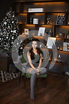 Fashionable and sexy young woman with slim perfect body sits near Christmas tree in the decorated interior. Beautiful