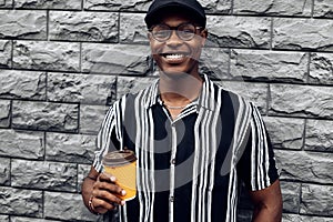 Fashionable smiling african american man in hat, man outdoors with takeaway coffee cup, over gray wall background