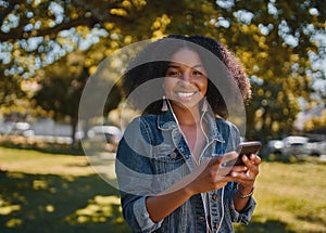 Fashionable smiling portrait of a young african american woman listening to music on smartphone in park looking at