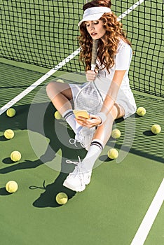 fashionable sportswoman in white sportswear with tennis racket taking selfie on smartphone at net