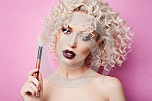 Fashionable and stylish model girl with blue eyes and curly blond hair, with professional bright makeup posing with makeup brush i