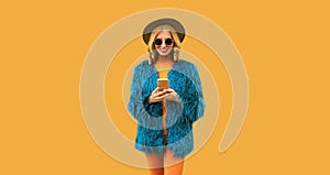 Fashionable stylish young woman with mobile phone wearing blue faux fur, round hat on yellow studio background