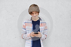 Fashionable teenage boy wearing shirt, holding cell phone in hands, messaging with friends or playing games online using free inte