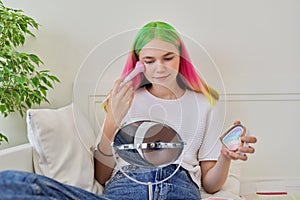 Fashionable teenage girl at home on bed with make-up mirror, holding brush and highlighter