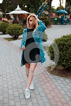 Fashionable urban cute young woman in a black stylish dress in a trendy long denim jacket in white sneakers posing in a park