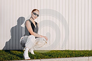 A fashionable woman with bun hair and black sunglasses on her eyes, wearing a black vest, white baggy pants and white high heeled