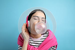 Fashionable woman with headphones blowing bubblegum on light blue background