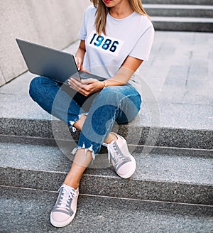 Fashionable woman sitting on stairs with laptop, wearing ripped jeans,white T-shirtand sneakers. fashion blogger outfit details