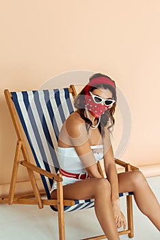 Fashionable woman in sunglasses, mask and bathing suit sitting on deck chair