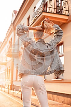 Fashionable woman in a trendy blue denim jacket in white vintage jeans straightens her hair in the city at sunset. Back view
