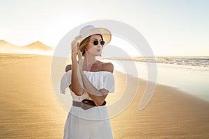 Fashionable woman in white dress and summer hat posing on the sandy beautiful beach