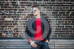 Fashionable young adult stylish man posing outdoors wearing red pulower, jeans, cotton jacket and sunglasses.