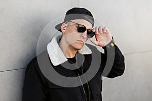 Fashionable young American hipster man in stylish sunglasses in a stylish black baseball cap in a black trendy jacket poses