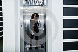 Fashionable young beautiful african american business woman with afro hairstyle wear in elegant black, stand at elevator with