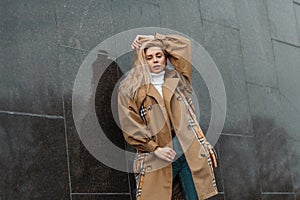 Fashionable young beautiful blond woman with trendy hairstyle in a stylish beige coat posing near a black modern wall in the city