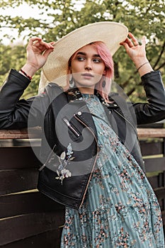 Fashionable young beautiful woman with pink hair in vintage straw hat in stylish black leather jacket in trendy blue dress near