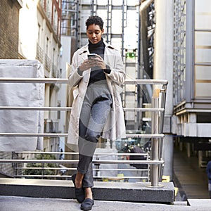 Fashionable young black woman standing in the city leaning on a hand rail using her smartphone, full length, low angle