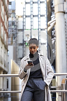 Fashionable young black woman standing in the city leaning on a hand rail using her smartphone, vertical