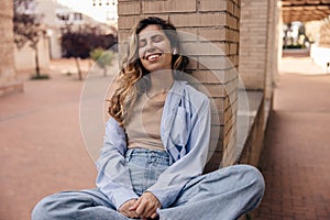 Fashionable young caucasian woman smiles teeth, uses headphones sitting on street during day.