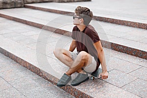 Fashionable young hipster man in black sunglasses in a vintage t-shirt in trendy shorts in sneakers resting while sitting