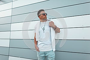Fashionable young hipster man with trendy hairstyle in sunglasses in jeans in a white T-shirt posing near modern building on the