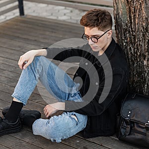 Fashionable young hipster man in vintage glasses in trendy denim casual wear in sneakers with leather backpack is resting near