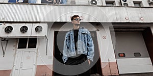 Fashionable young hipster man in youth stylish clothes in trendy sunglasses stands near a vintage white building in the city on a