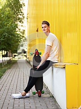 Fashionable young hipster sits with a skateboard on a blurred natural background. Active lifestyle concept. Copy space.