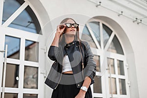 Fashionable young hipster woman in straightens trendy sunglasses. Stylish urban girl model in seasonal clothes posing in the city