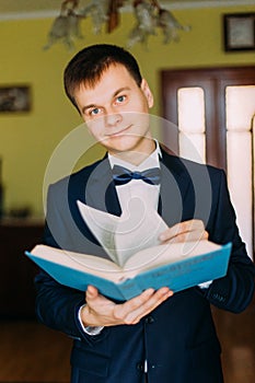 Fashionable young man with bow-tie holding a book and looking at the camera. Hotel room in the background