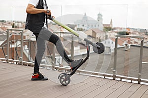 Fashionable young man in a denim vest in a T-shirt in gray jeans in sneakers posing with an electric scooter on a summer terrace.