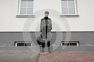 Fashionable young man in a gray elegant plaid coat with a leather stylish black backpack in autumn trendy boots stands near a