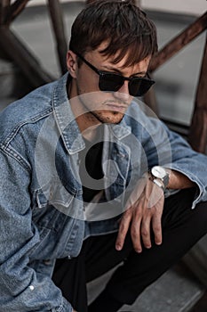 Fashionable young man hipster in stylish denim clothes trendy in a black sunglasses sits on a vintage staircase in the city