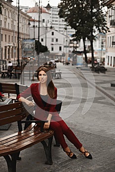 Fashionable and young pretty woman, in a cherry smart suit, sitting on the bench and relaxing outdoors. Stylish brunette