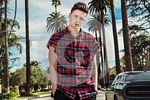 Fashionable young man wearing street style outfit standing on city street near car and palm at hot summer day photo