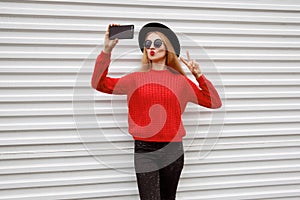 Fashionable young woman blowing red lips sends sweet air kiss taking selfie picture by phone in knitted sweater