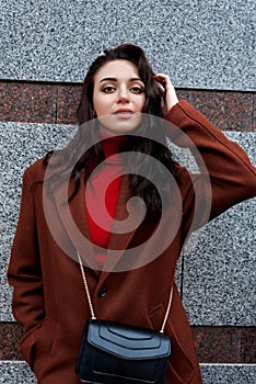 fashionable young woman in a brown coat poses against the wall