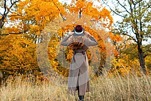 Fashionable young woman in an elegant long coat in a stylish hat stands among the dry grass and enjoys the autumn scenery in the