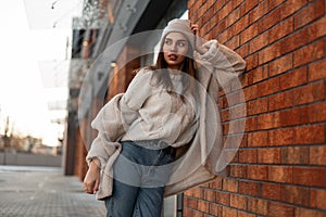 Fashionable young woman model in a stylish hat in a trendy coat in blue jeans in a vintage sweater is resting near a vintage brick