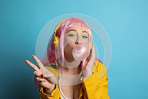 Fashionable young woman in pink wig with headphones blowing bubblegum on yellow background