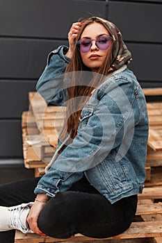 Fashionable young woman in purple glasses in stylish clothes puts military hood on head. American trendy hipster girl in youth