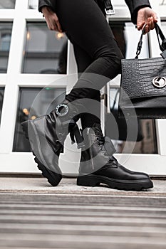 Fashionable young woman stands on the stone steps in the city in black trendy leather boots in vintage jeans with a bag. Close up