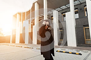 Fashionable young woman in stylish black blazer walks near building in city beautiful at sunset. Lovely gorgeous girl travels and