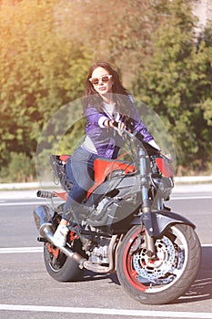 Fashionable young woman with sunglasses, lilac short windbreaker and jeans with a beautiful body on a red sports bike