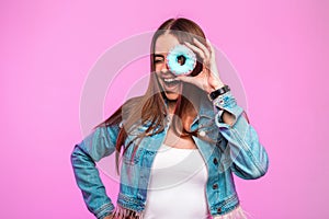 Fashionable young woman trendy in jeans wear covers eyes with sweet donut near vintage pink wall in the room. Studio creative