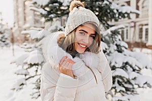 Fashionable young woman in white knitted hat smiling friendly to camera on street full with snow. Amazing european girl