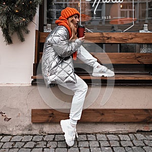 Fashionable young woman in youth outerwear in stylish sneakers in a knitted hat with an orange scarf is resting on a wooden bench