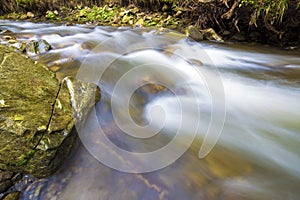 Fast flowing through wild green forest river with crystal clear smooth silky water falling from big wet stones in beautiful