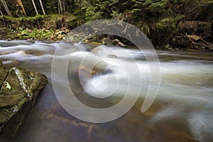 Fast flowing through wild green forest river with crystal clear smooth silky water falling from big wet stones in beautiful