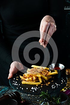 Fast food, french fries. Potatoes with bacon on a plate in the hands of a chef, on rustic wooden background.